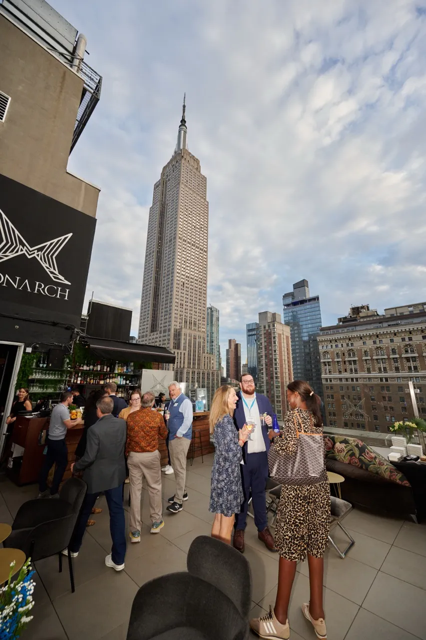 People socializing on a rooftop bar with the Empire State Building and city skyline in the background during daytime.