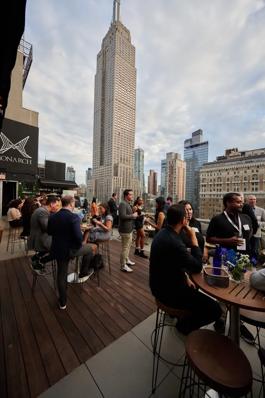People socializing on a rooftop terrace with the Empire State Building and city skyline in the background during daytime.