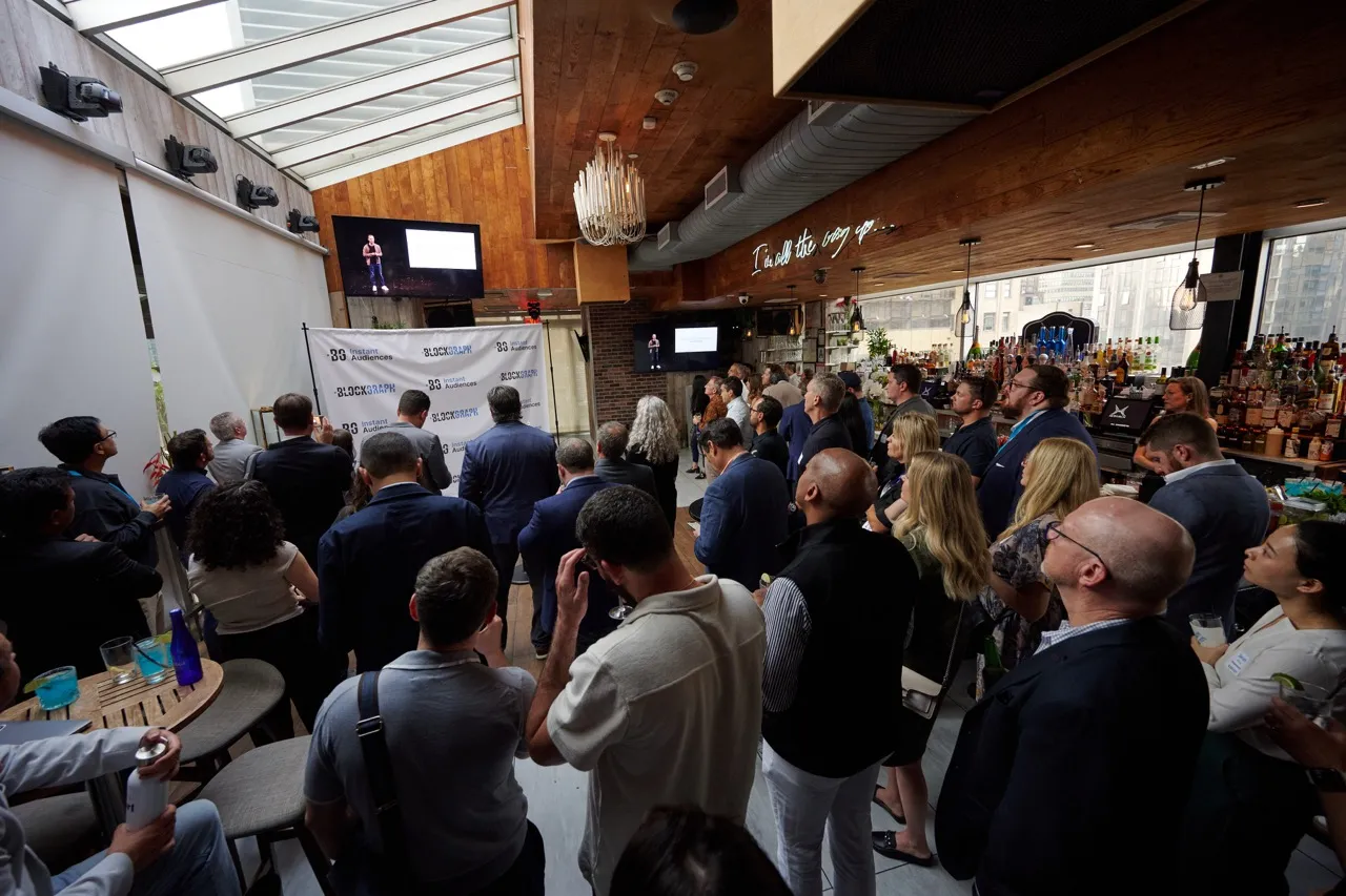 Audience of professionals watching a presentation on screens in a modern bar venue with wooden ceiling and large windows.