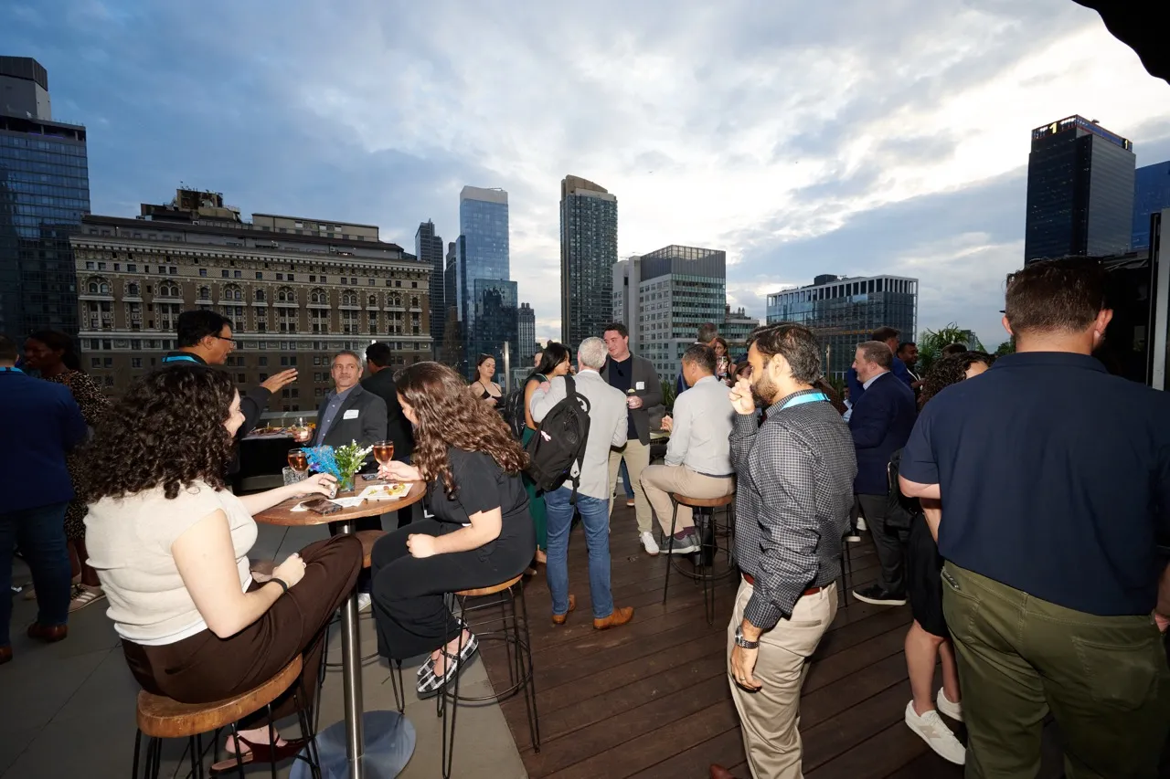 People socializing at an outdoor rooftop event with city buildings in the background under a cloudy sky.