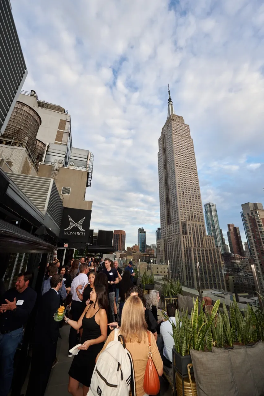 People socializing on a rooftop terrace with the Empire State Building and city skyline in the background under a partly cloudy sky.