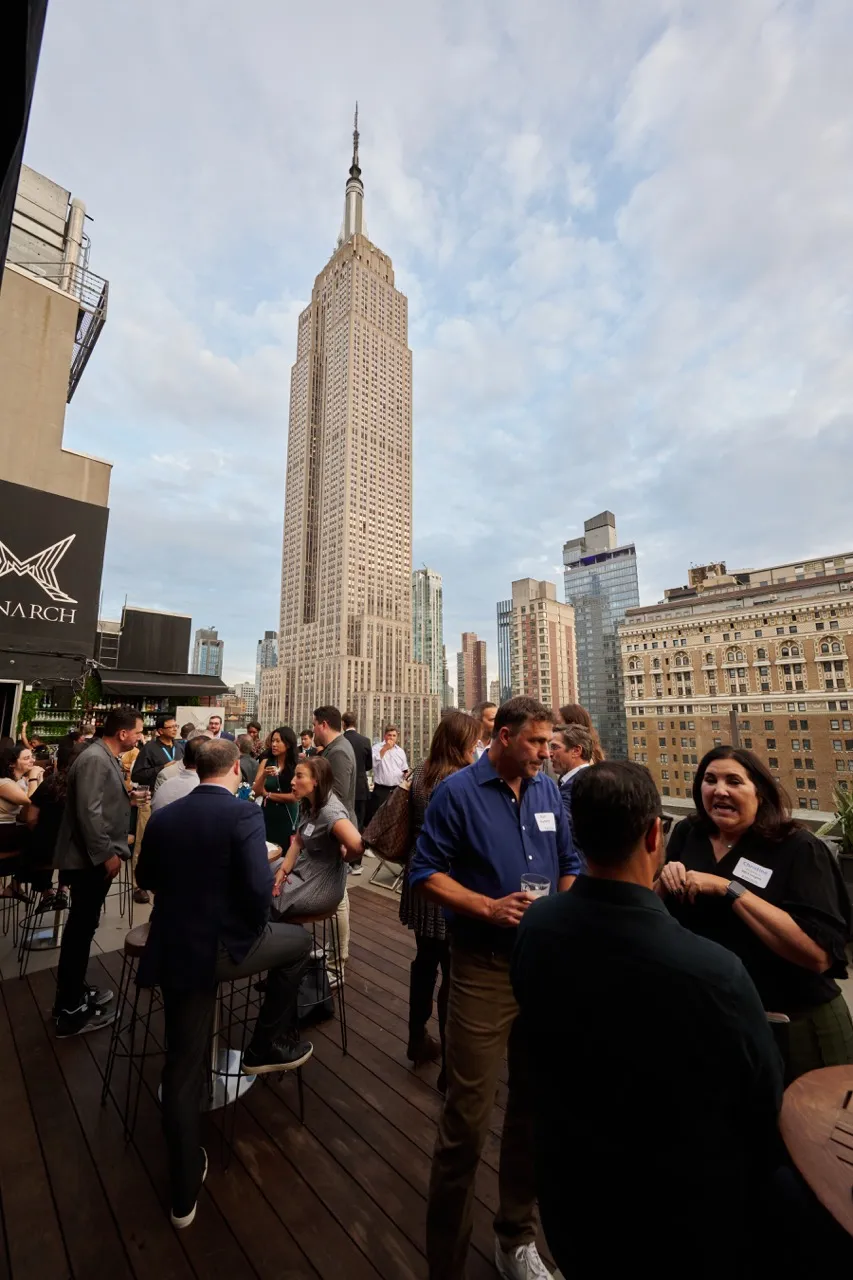 People socializing on a wooden rooftop terrace with the Empire State Building and city skyline in the background under a partly cloudy sky.