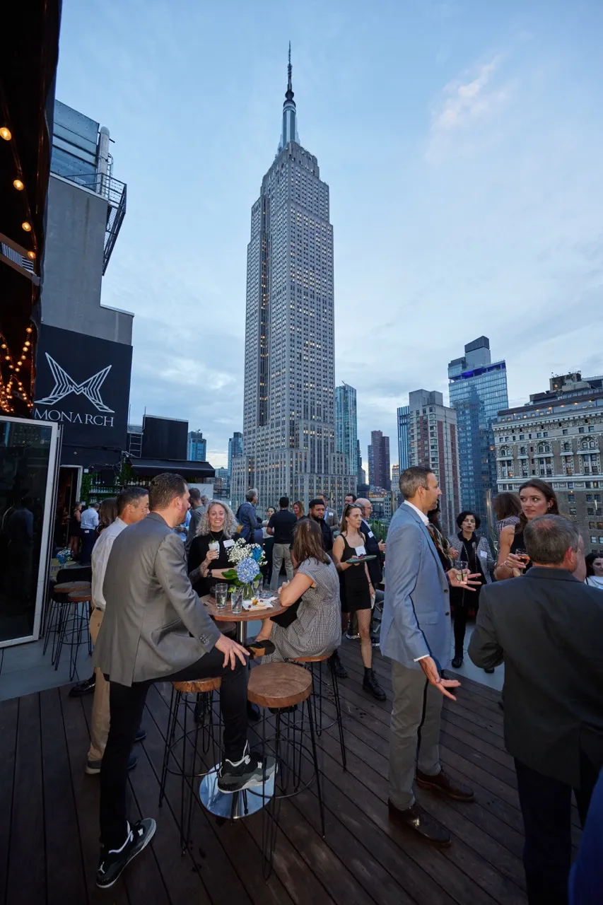 People socializing on a rooftop deck with the Empire State Building and other skyscrapers in the background at dusk.