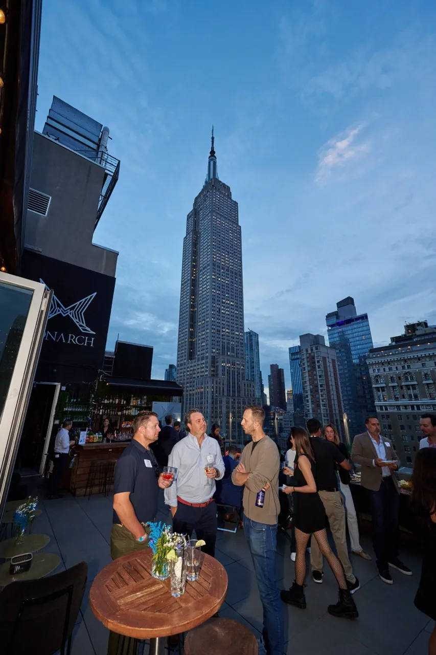 People socializing with drinks on a rooftop terrace at dusk with the Empire State Building in the background.