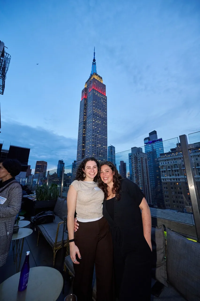 Two smiling women posing on a rooftop terrace with the Empire State Building illuminated in red, yellow, and blue behind them at dusk.