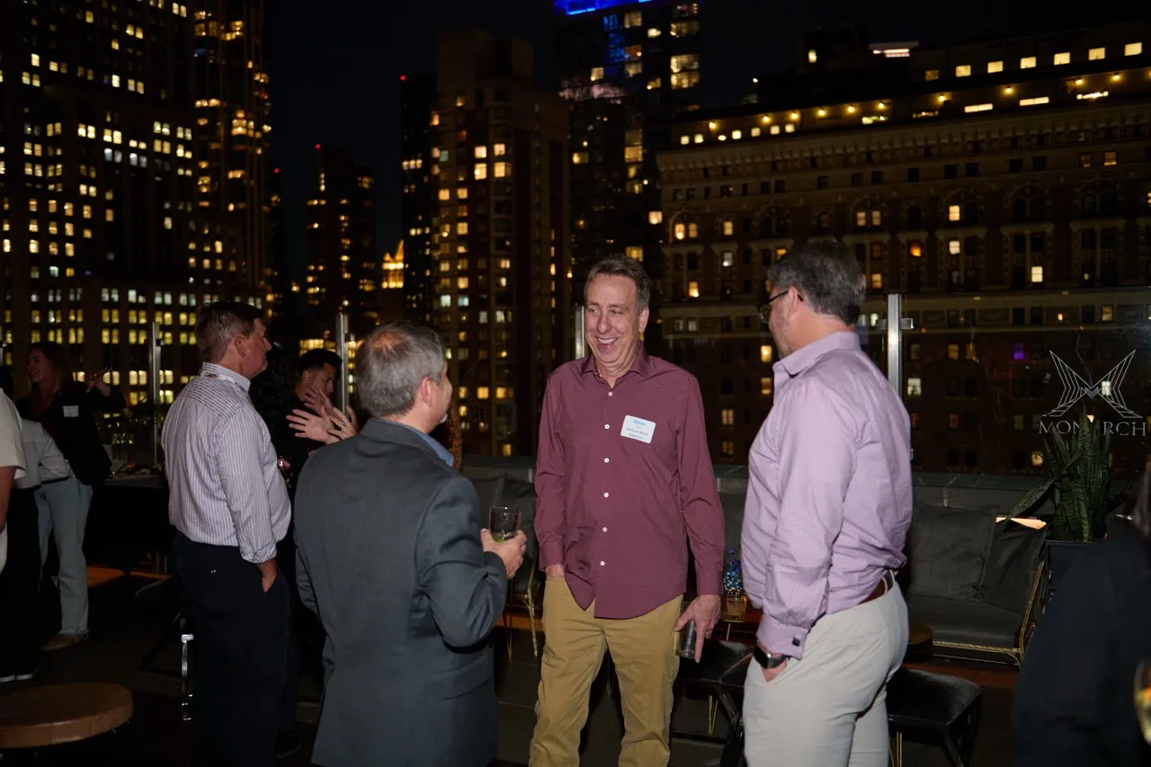 Group of men socializing at an outdoor rooftop event at night with city lights in the background.