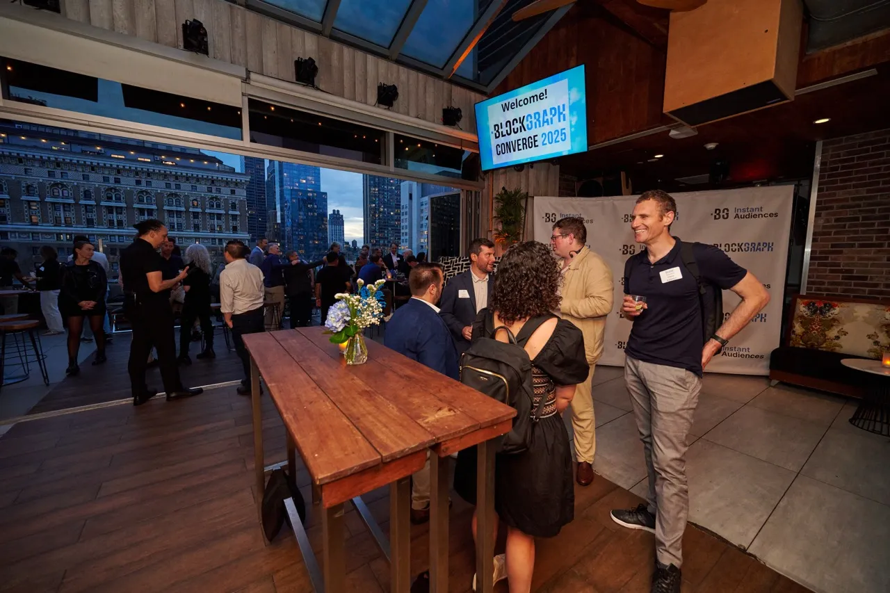 People socially mingling at an indoor rooftop event with city buildings visible outside, a wooden table with a flower vase in the foreground, and a screen reading 'Welcome! BLOCKGRAPH CONVERGE 2025' overhead.