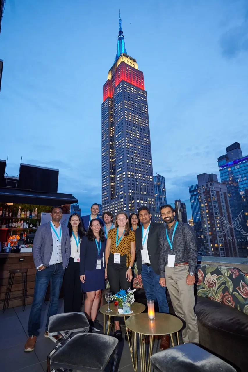Group of nine people posing on a rooftop with the Empire State Building lit in red, white, and blue in the background during dusk.