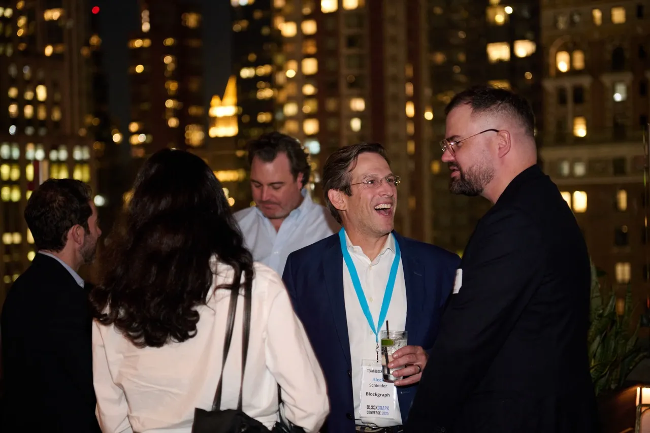 Group of people socializing at night with city lights in the background, one man holding a drink and wearing a conference badge.