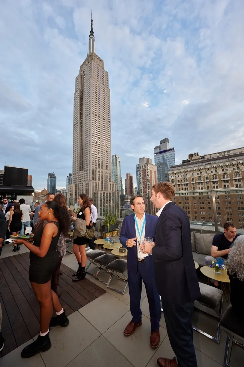 People socializing on a rooftop terrace with the Empire State Building and other skyscrapers in the background under a cloudy sky.
