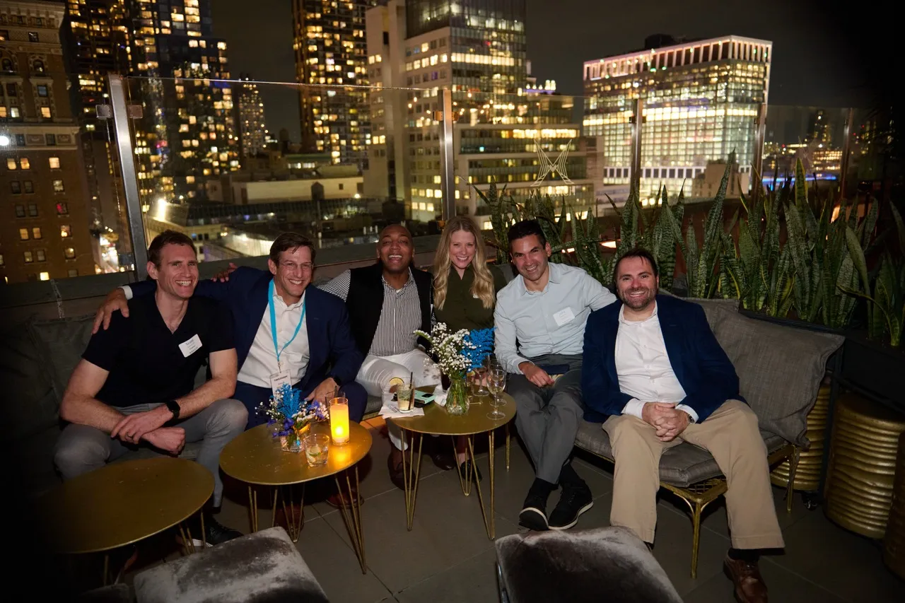 Six people sitting closely on a rooftop lounge with city lights and buildings in the background at night.