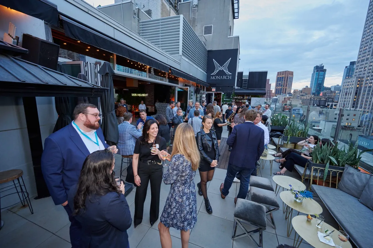 Group of people socializing on a modern rooftop patio with city skyline in the background during a casual evening event.