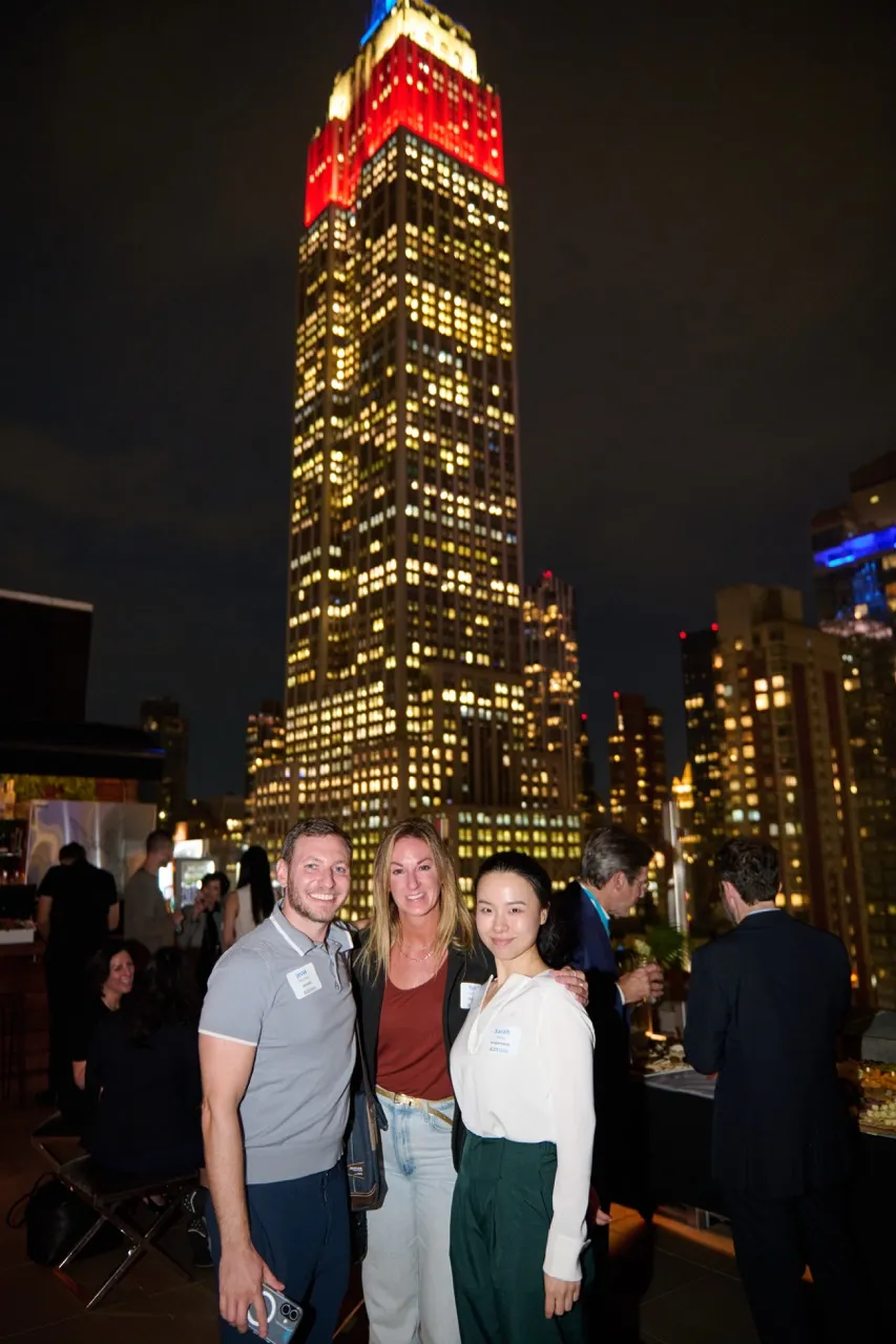 Three people smiling and posing on a rooftop at night with the illuminated Empire State Building in the background.