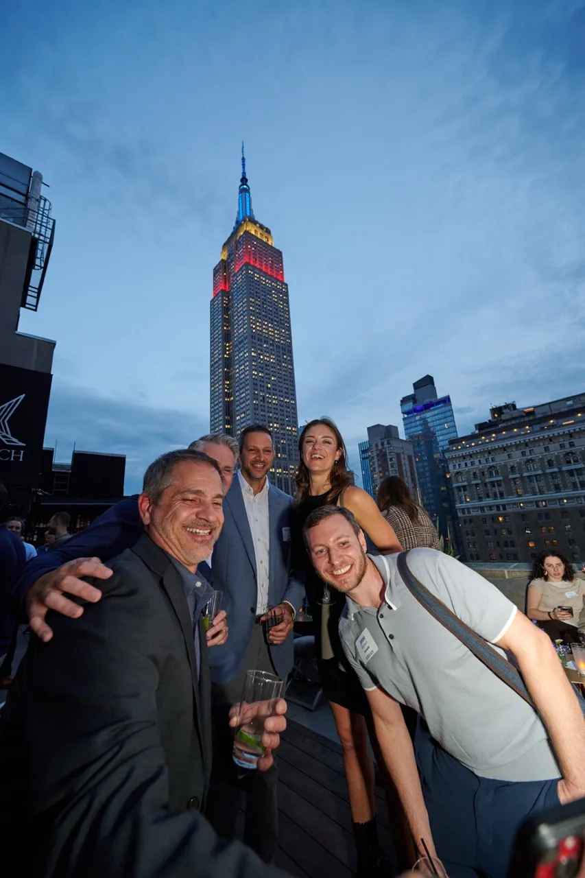 Group of five smiling people posing for a photo on a rooftop with the Empire State Building lit in red, yellow, and blue in the background at dusk.
