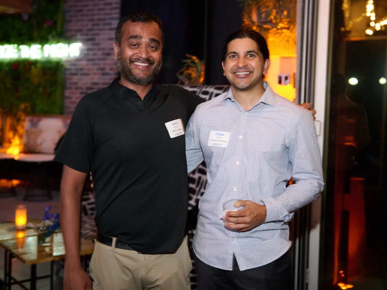 Two men smiling indoors at an event with name tags, one wearing a black shirt and beige pants, the other in a light checkered shirt holding a drink.