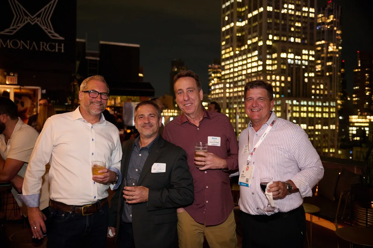 Four men smiling and holding drinks at a nighttime rooftop event with city lights in the background.