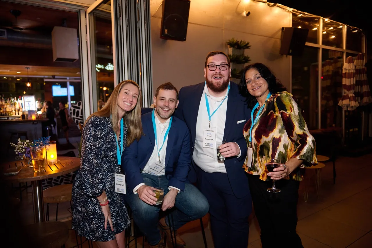 Four people smiling and posing together indoors at a social event, wearing name badges and holding drinks.
