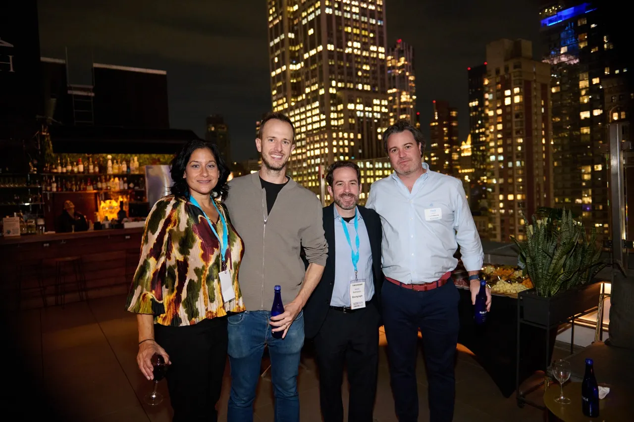 Four people standing on a rooftop at night with city skyline lights behind them, two men hold blue bottles and a woman holds a wine glass.