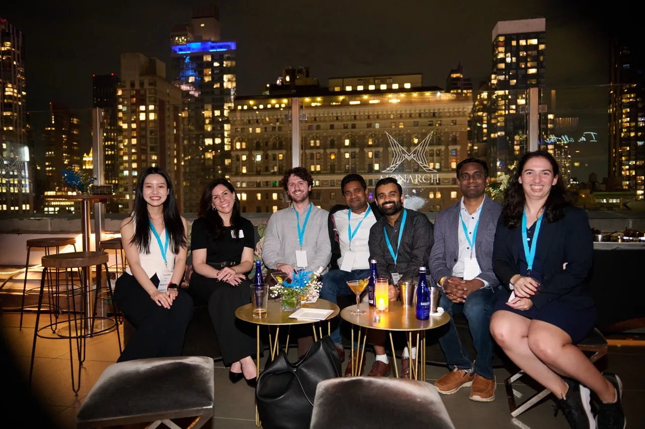 Seven people seated together on a night rooftop with city skyline in the background, smiling and wearing conference badges with blue lanyards.