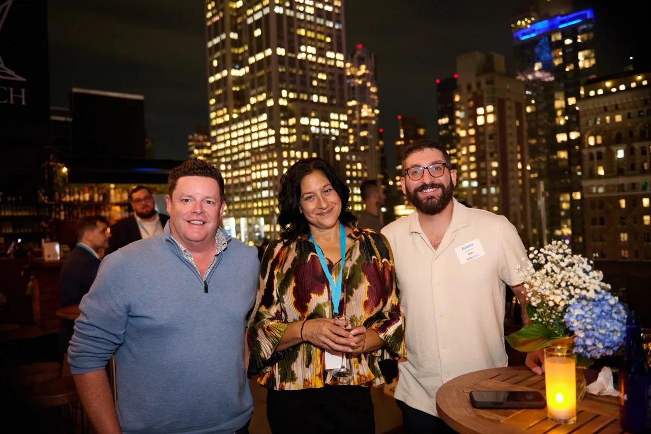 Three people smiling and posing at a night rooftop event with city lights in the background.