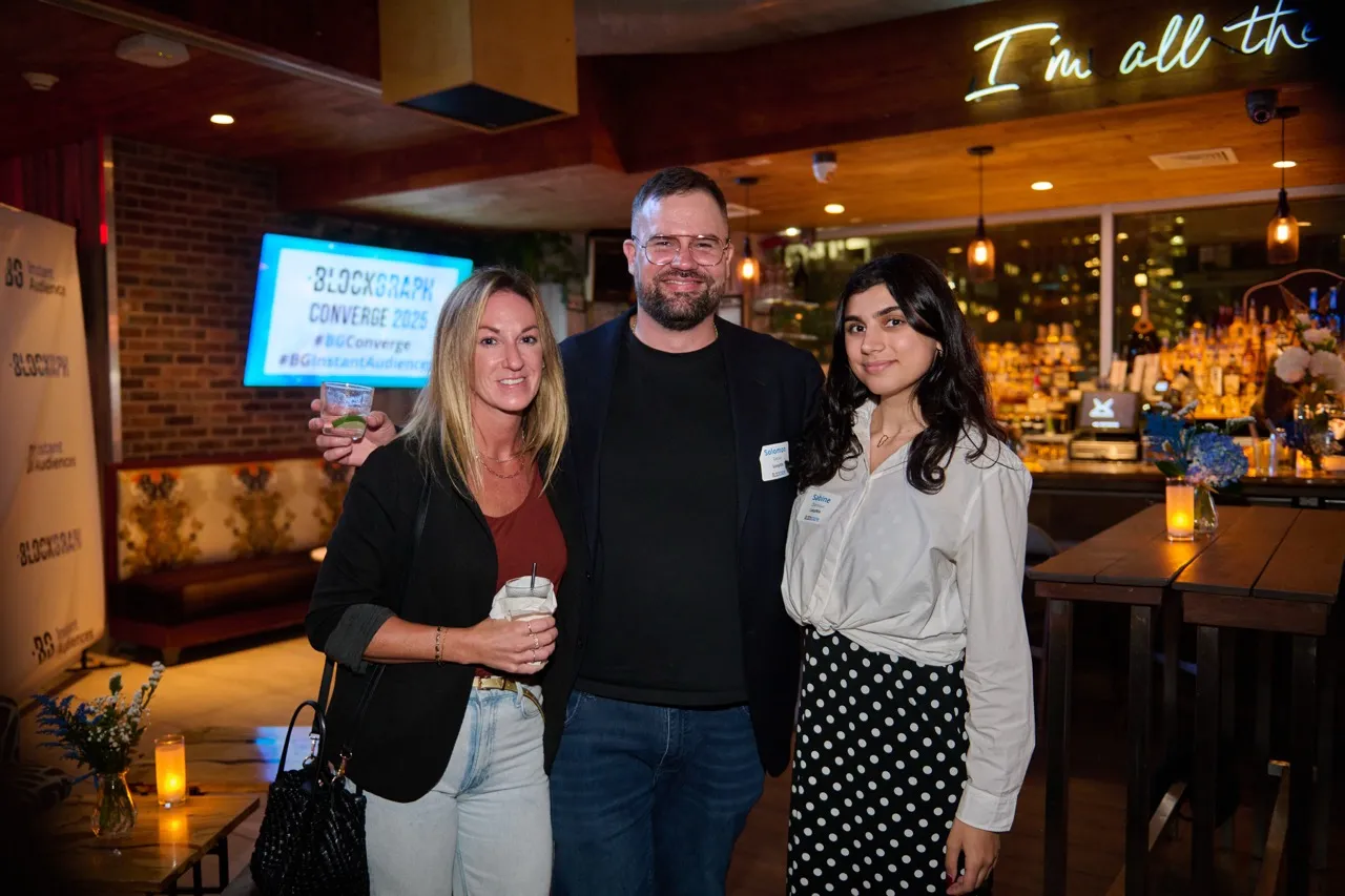 Three people smiling at a social event in a bar with cocktails, one man in the center and two women on either side.