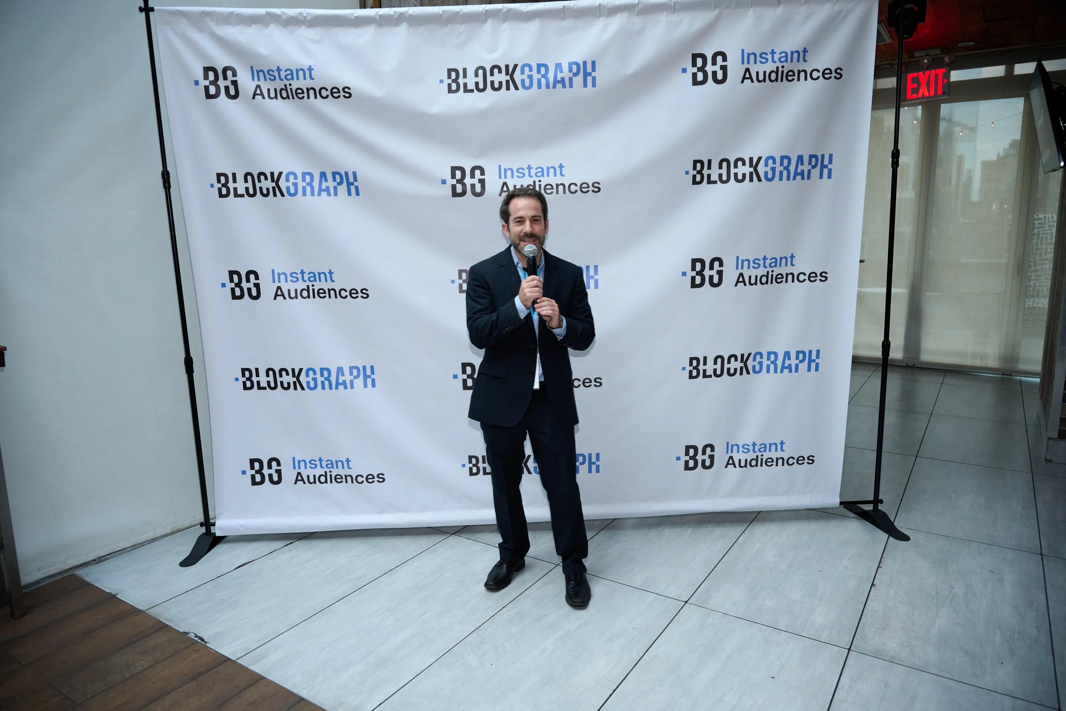 Man in a dark suit holding a microphone standing in front of a backdrop with Blockgraph and BG Instant Audiences logos.