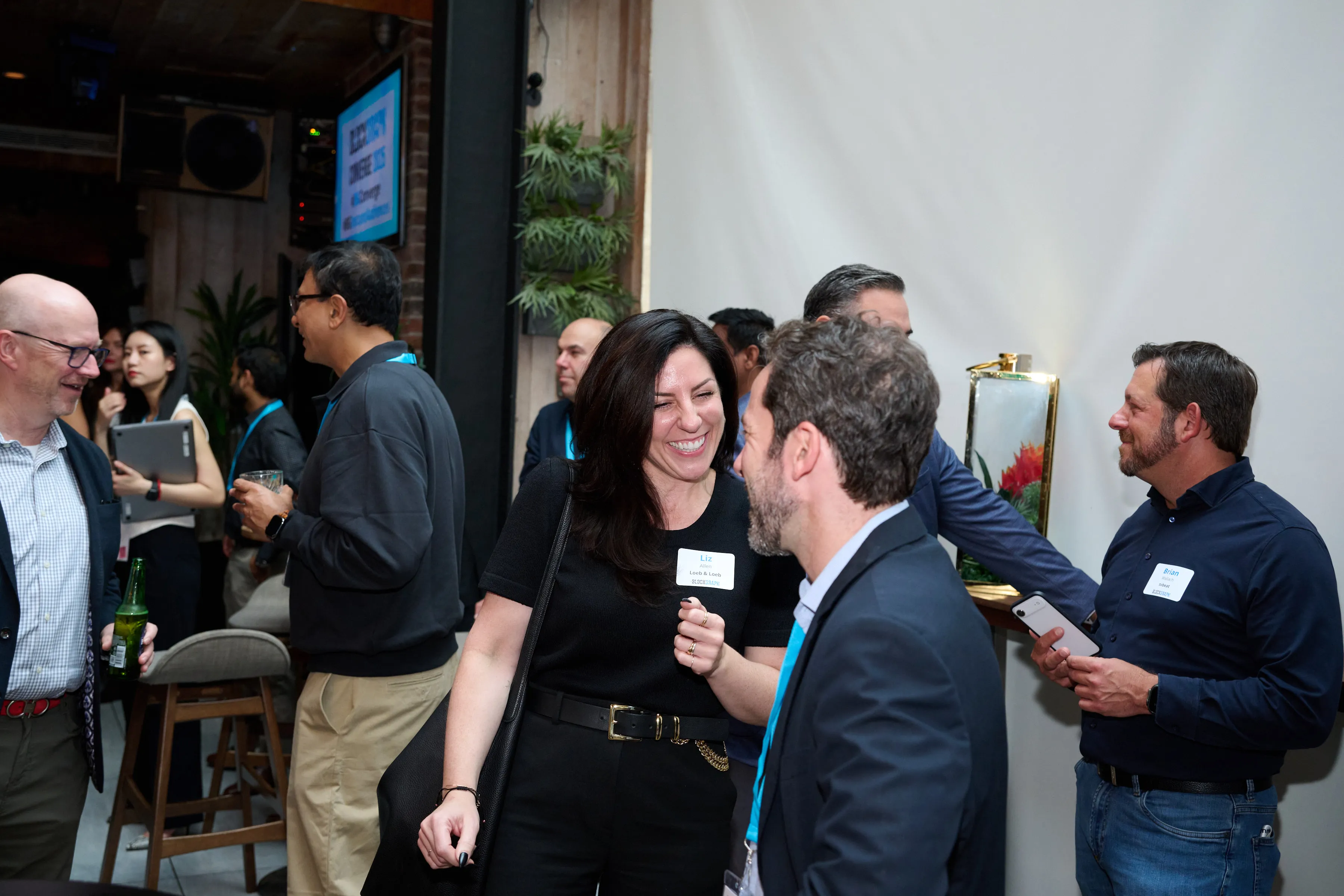 A group of people socializing at an indoor networking event, with a woman named Liz smiling and talking to a man in a suit.