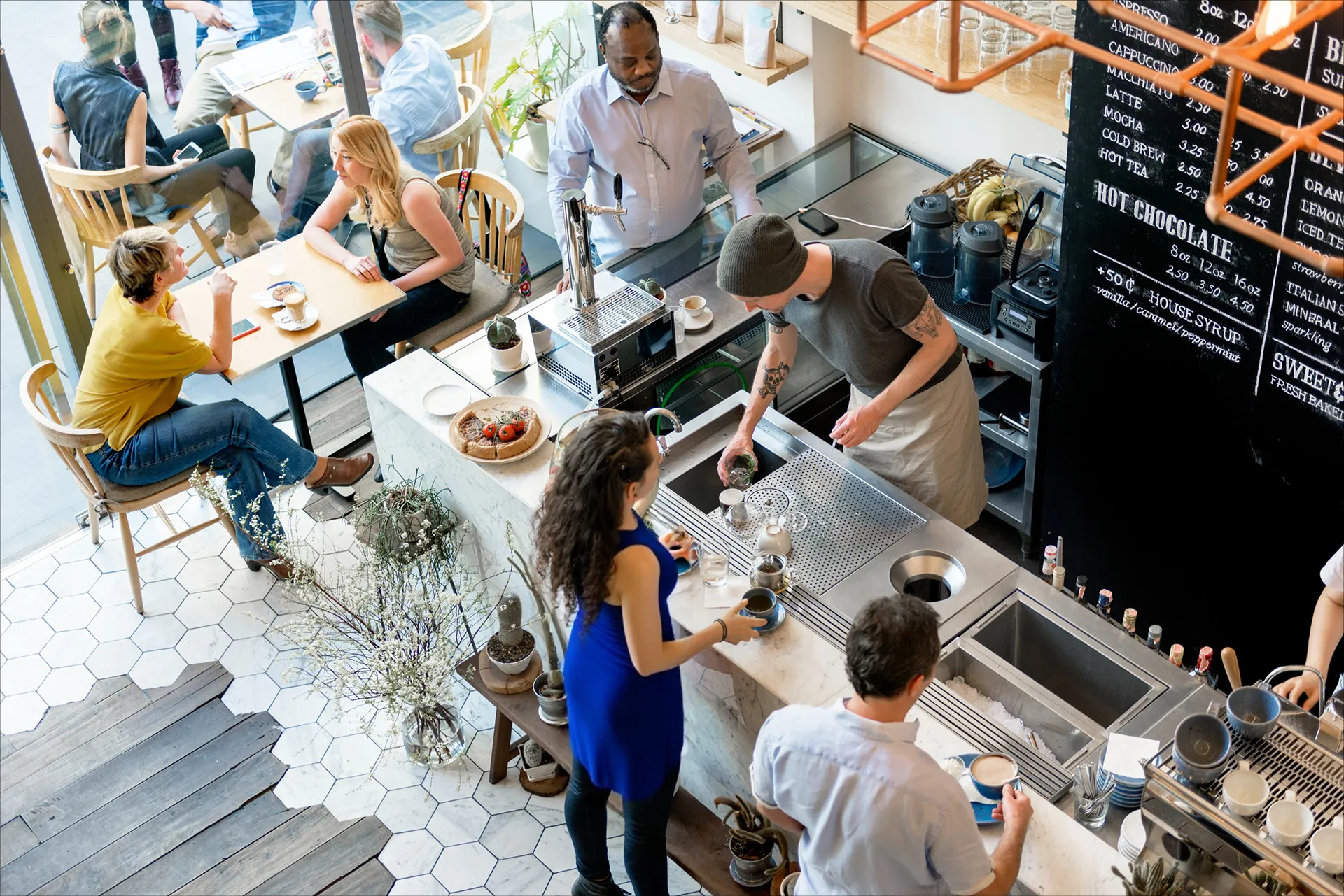 Busy café scene with customers sitting and chatting, and a barista preparing drinks behind the counter.