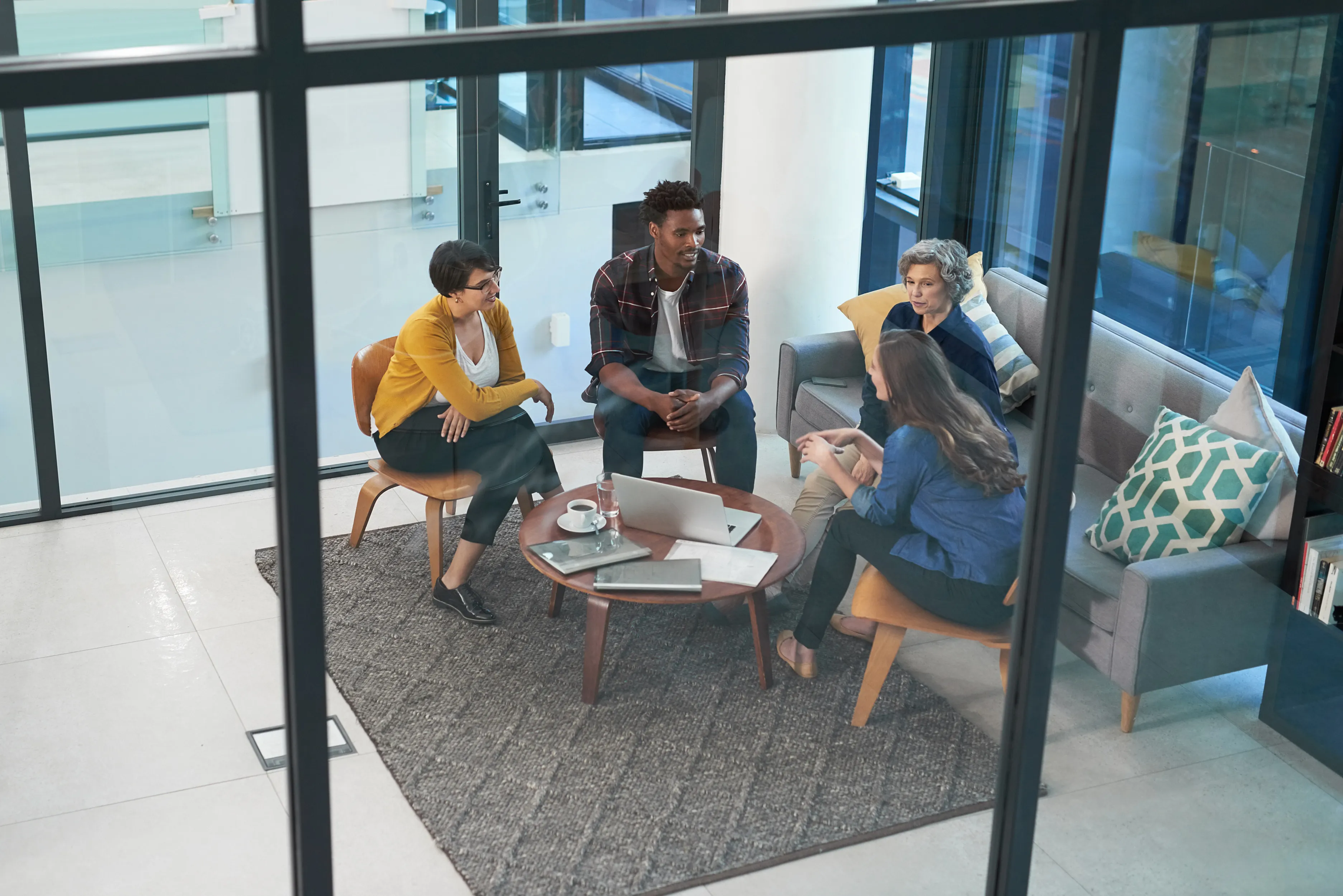 Four diverse colleagues having a discussion around a coffee table with a laptop and notebooks in a modern office lounge.