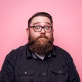 Man with glasses and a full beard wearing a dark button-up shirt against a pink background.