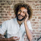 Smiling man with curly hair and beard talking on a mobile phone in front of a brick wall.