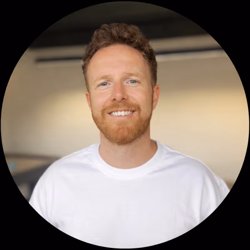 Smiling man with curly hair and red beard wearing a white shirt in a softly lit indoor setting.