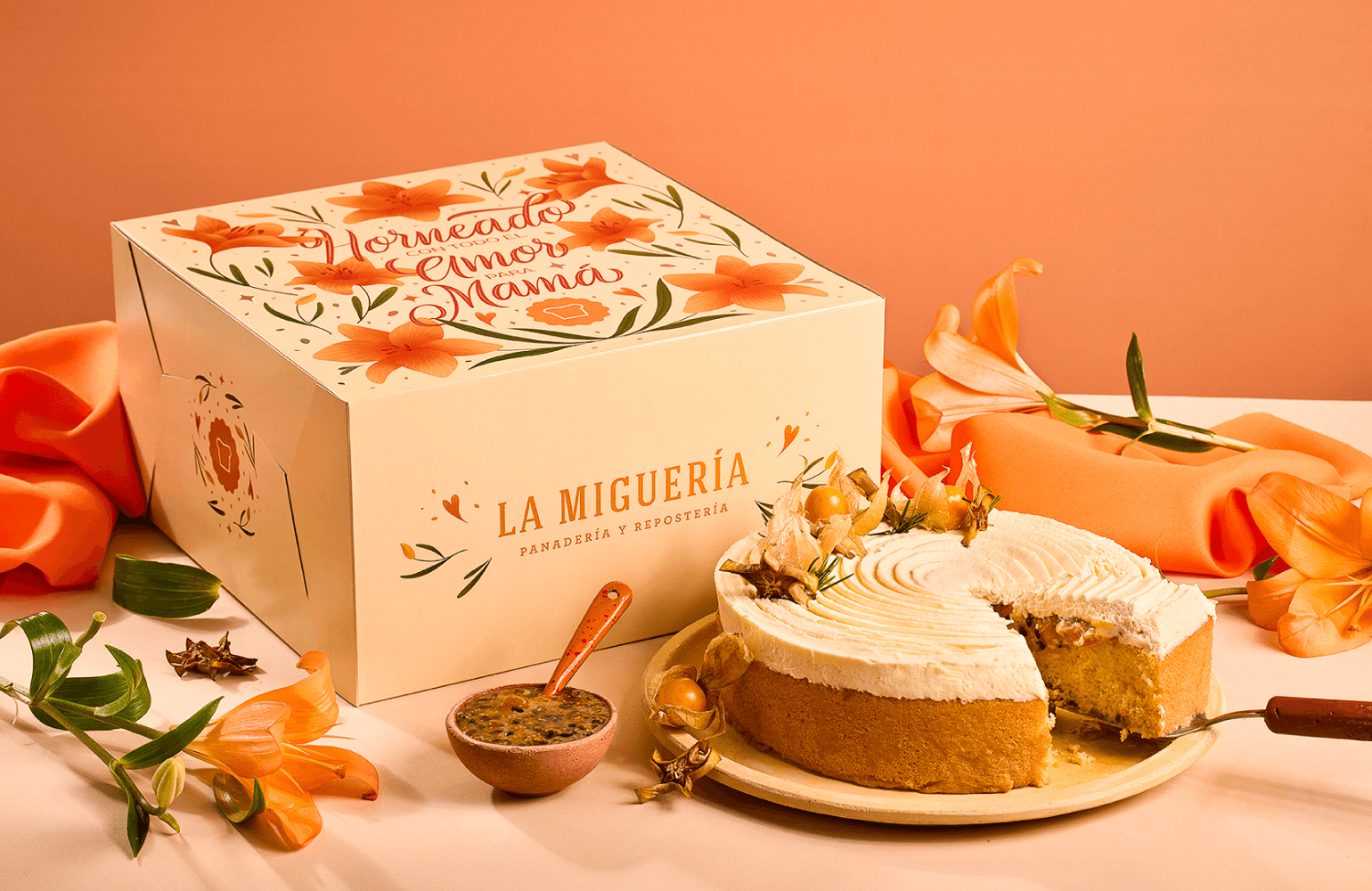 Torta artesanal con crema y uchuvas junto a caja de packaging naranja ilustrada con azucenas y lettering "Horneado con todo el amor para mamá" de La Miguería panadería y repostería colombiana fotografía de producto y diseño de packaging e ilustración por Intacto Studio estudio de branding en Medellín y Bogotá Colombia.