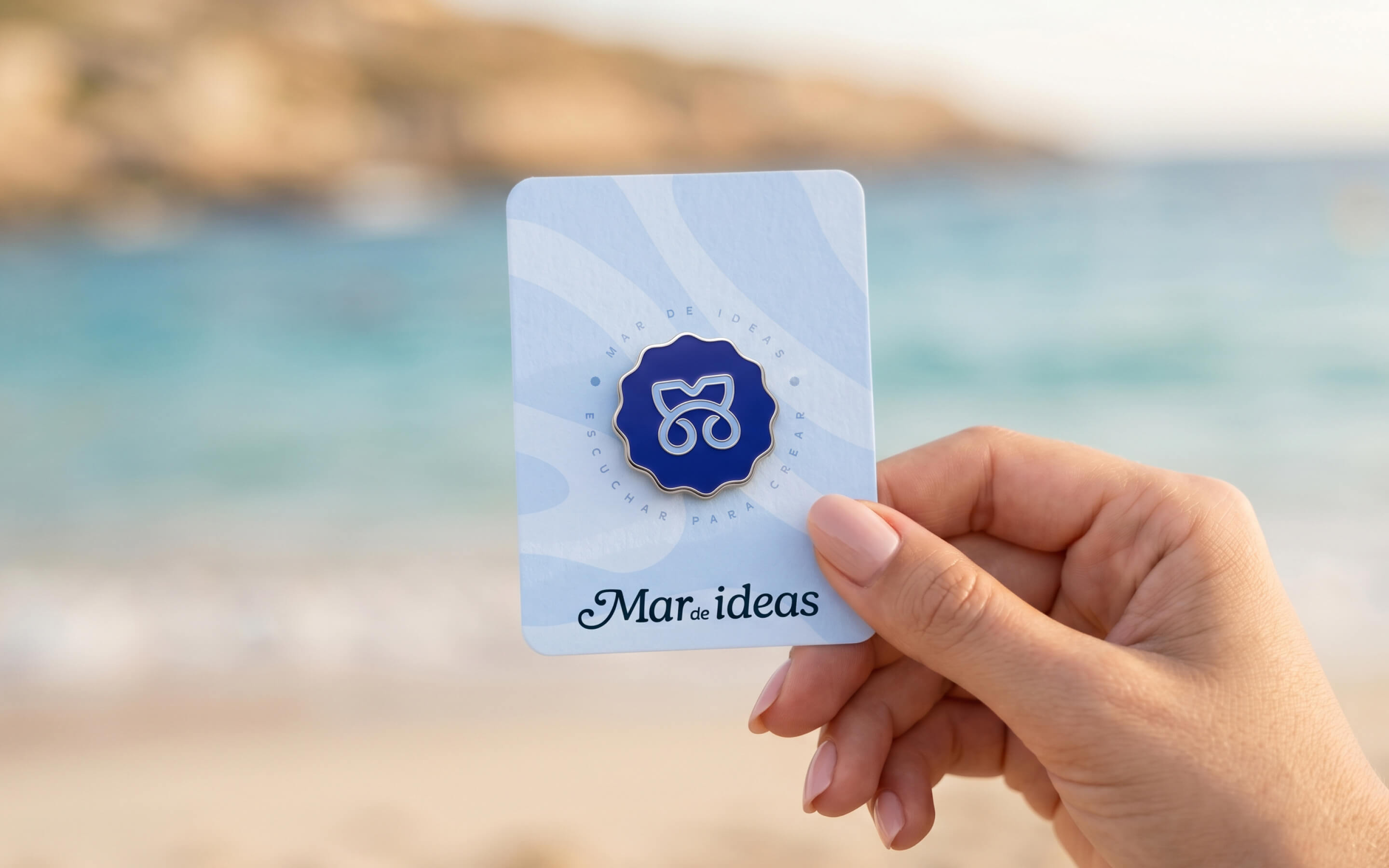 Tarjeta de marca de Mar de Ideas sostenida frente al mar, con pin del símbolo gráfico, patrones suaves y logotipo aplicado, como pieza de branding y material de identidad visual creado por Intacto Studio.