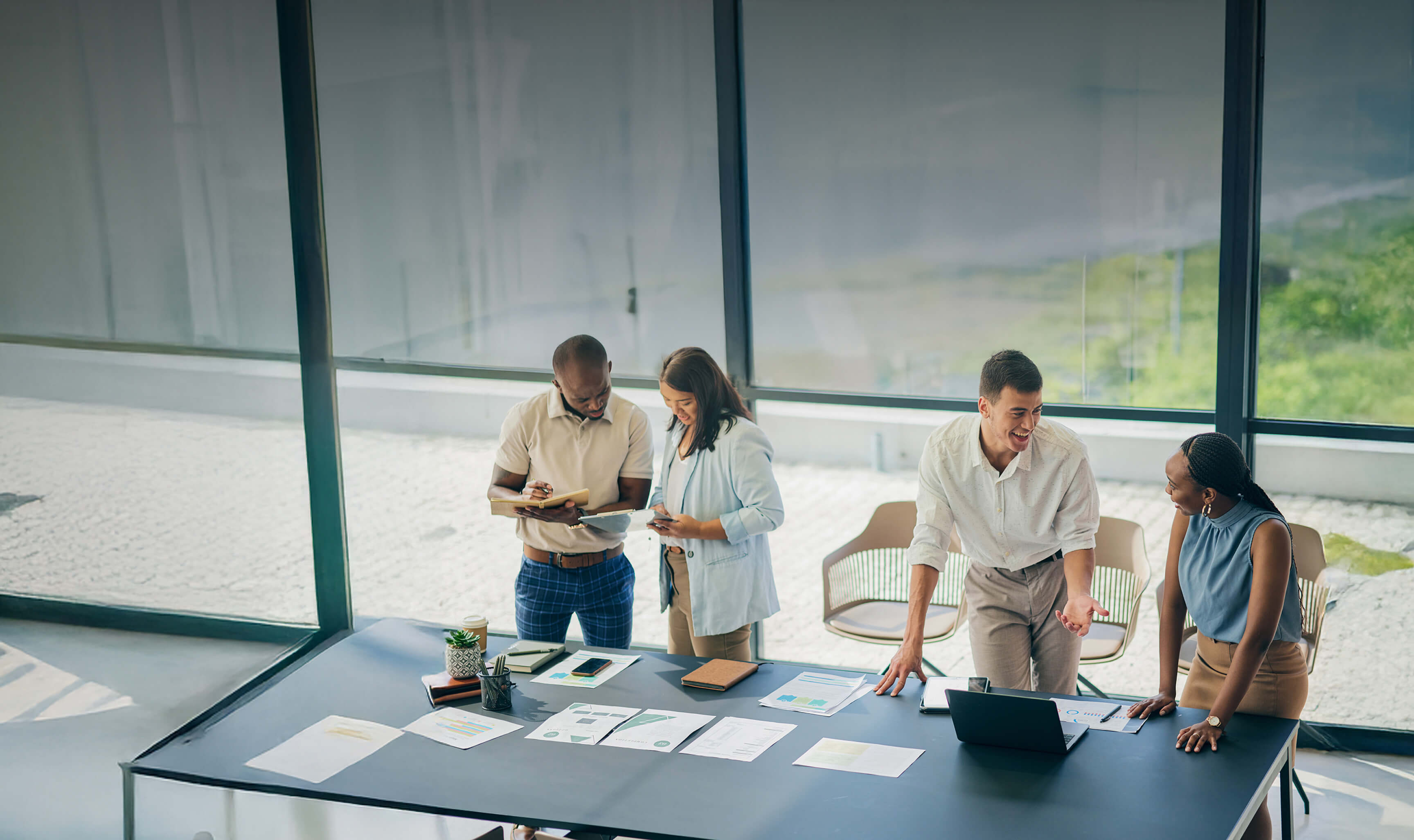 Four colleagues discussing work around a table with documents and a laptop in a bright office with large windows.