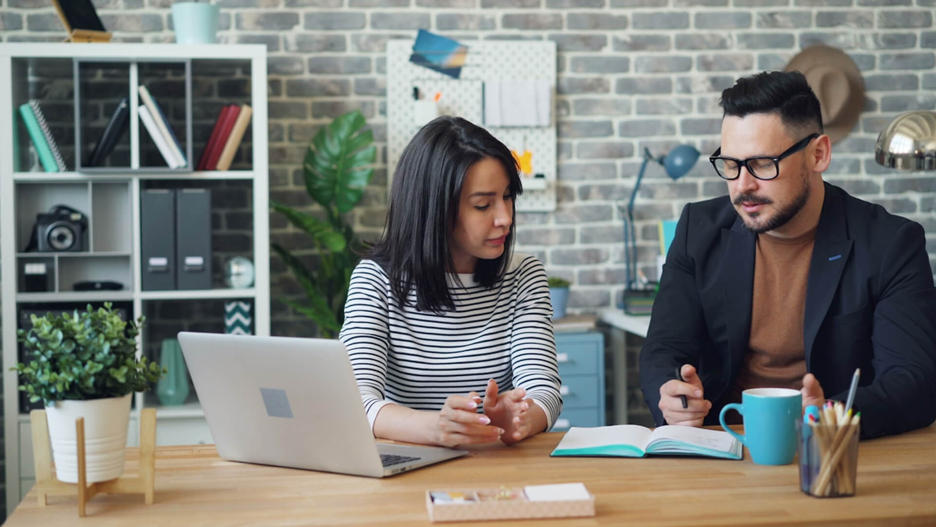 Two people sitting at a desk with a laptop, notebook, and coffee mug, discussing work in a modern office.