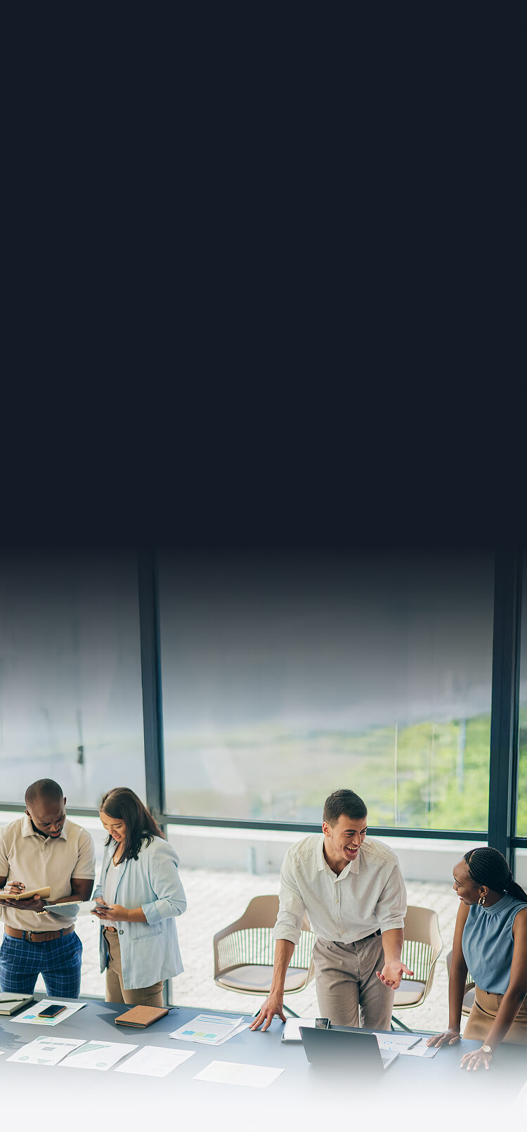 Four colleagues in business casual attire discussing documents and working around a table with laptops and papers in a modern office with large windows.