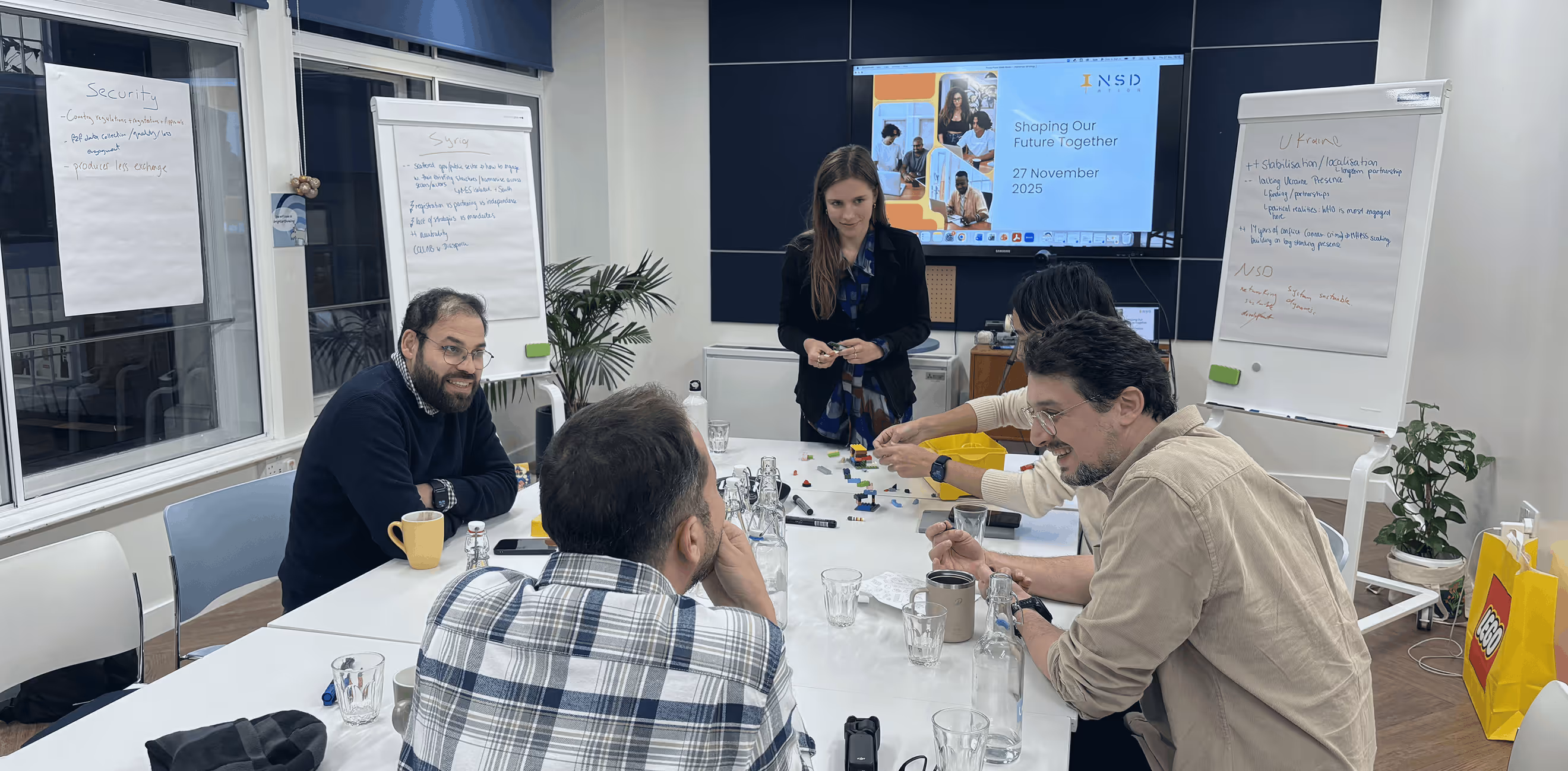 Five people in a meeting room around a white table with LEGO pieces, flip charts, and a presentation screen showing 'Shaping Our Future Together 27 November 2025'.