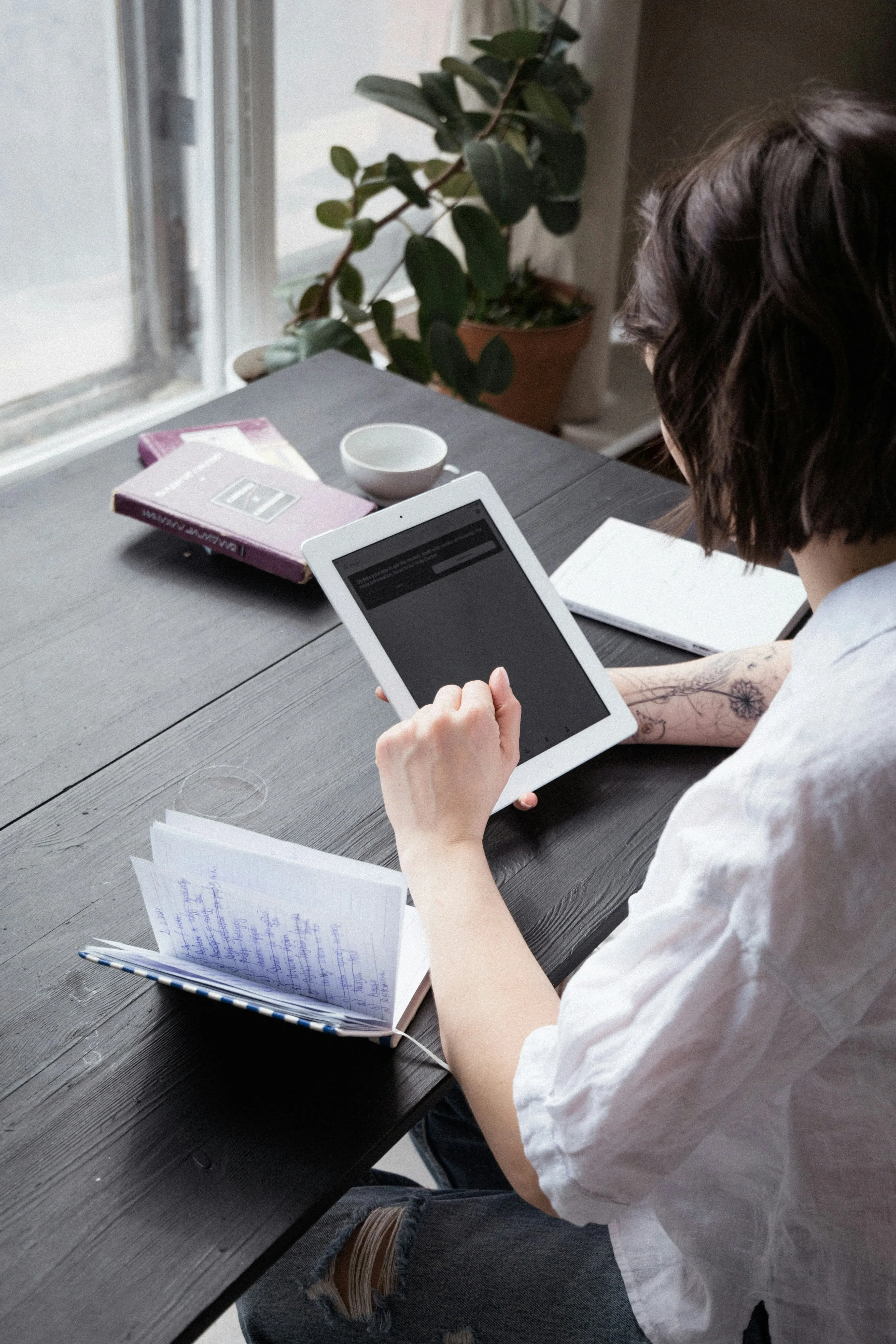 Person with a tattoo on their arm using a white tablet at a dark wooden table with books and a cup nearby.