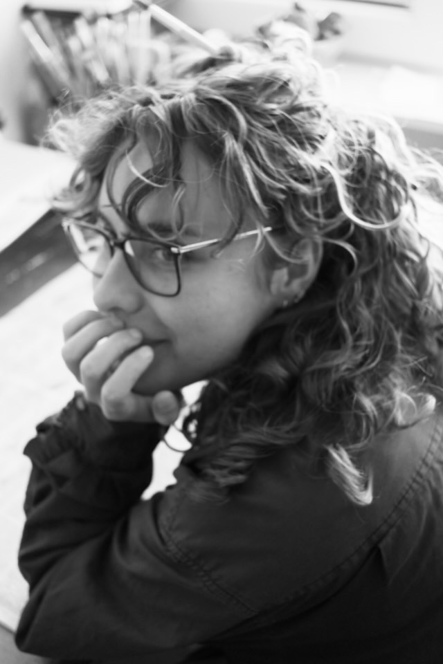 Black-and-white close-up of a person with curly hair and glasses resting their chin on their hand, looking sideways.