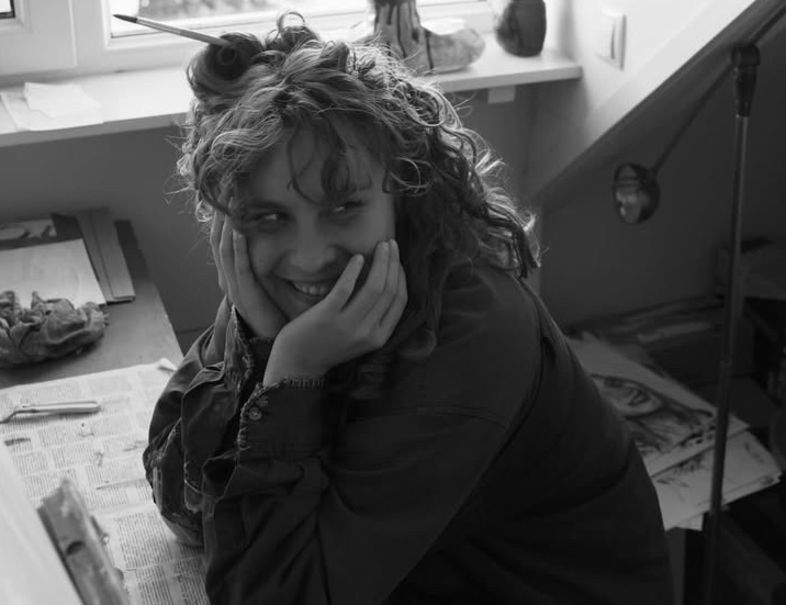 A smiling person with curly hair sits at a desk by a window, resting their face on hands with a pencil in their hair.