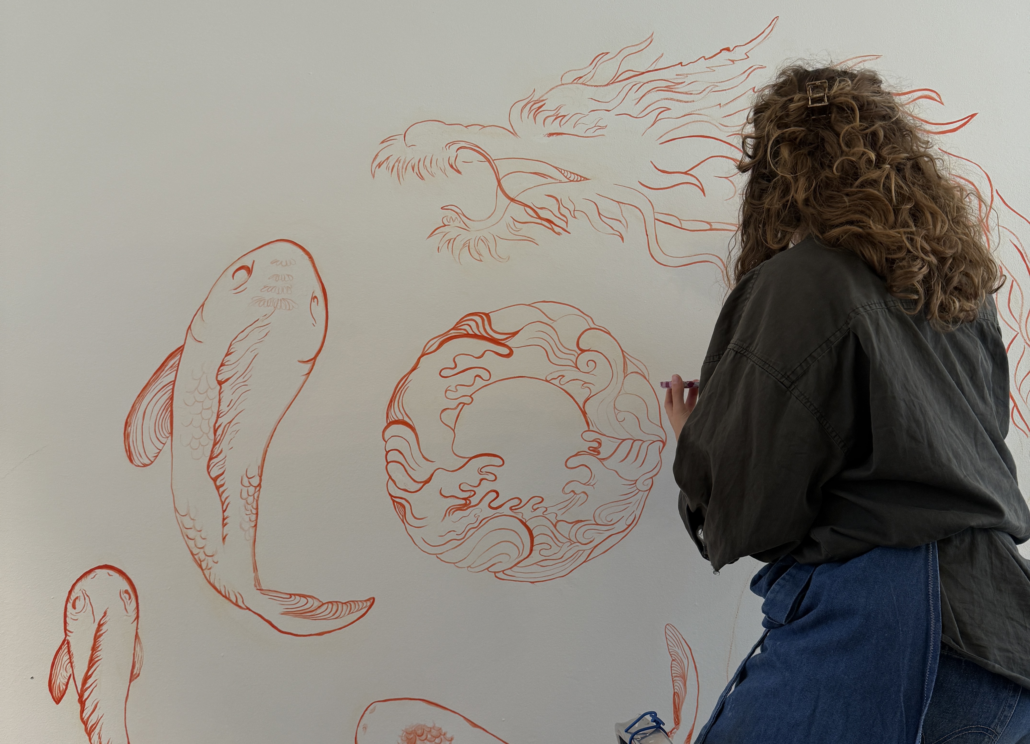 Woman with curly hair drawing intricate koi fish and dragon designs in orange on a white wall.