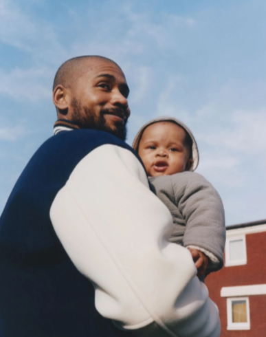 Man holding a baby outside with a blue sky and a red building in the background.