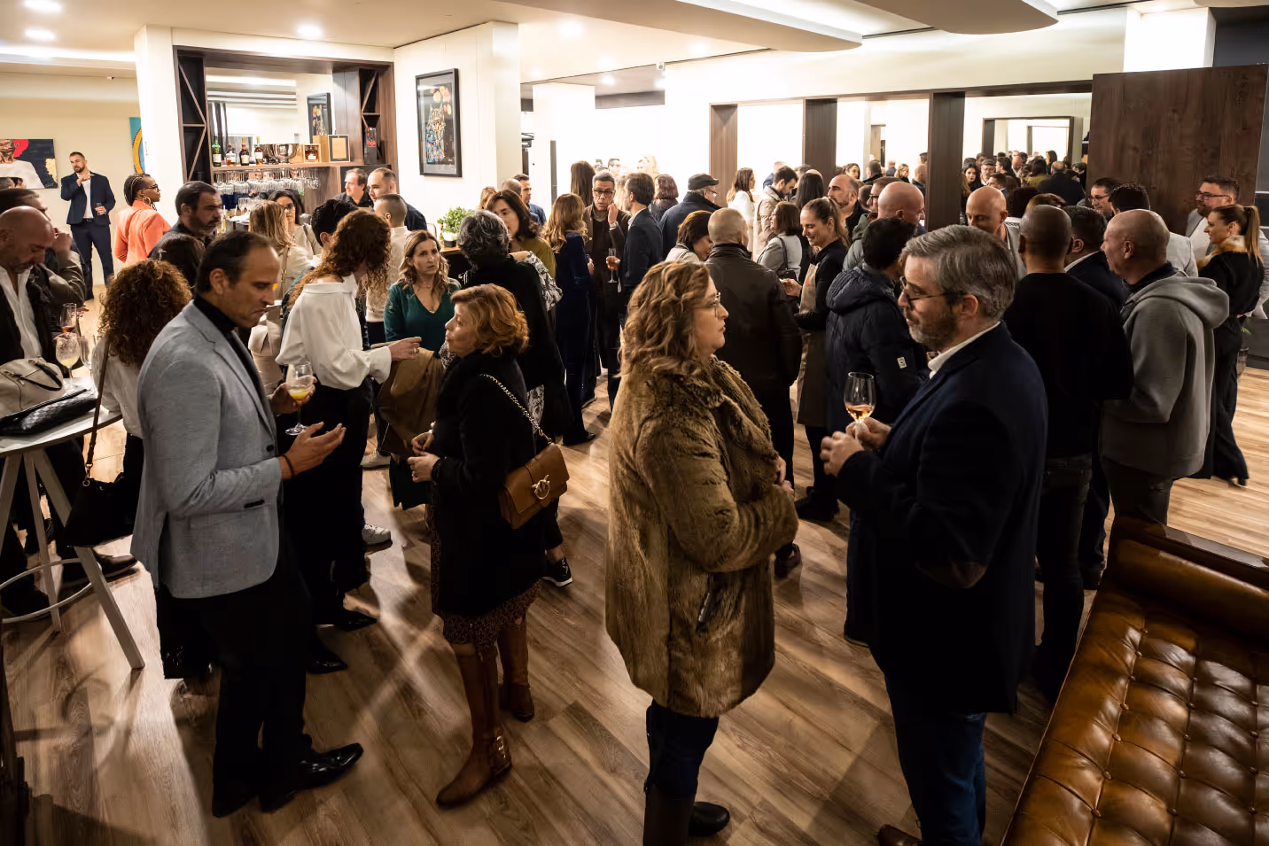 Multidão de pessoas socializando em evento interno com bebidas nas mãos e decoração moderna.
