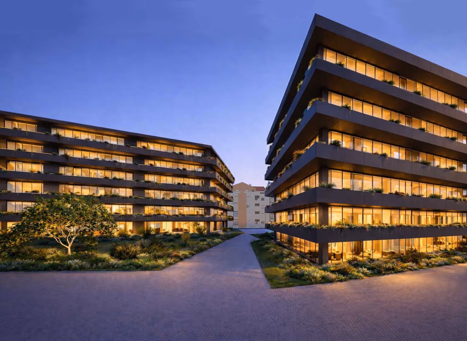 Dois edifícios modernos de escritórios com janelas iluminadas ao entardecer e área ajardinada entre eles.