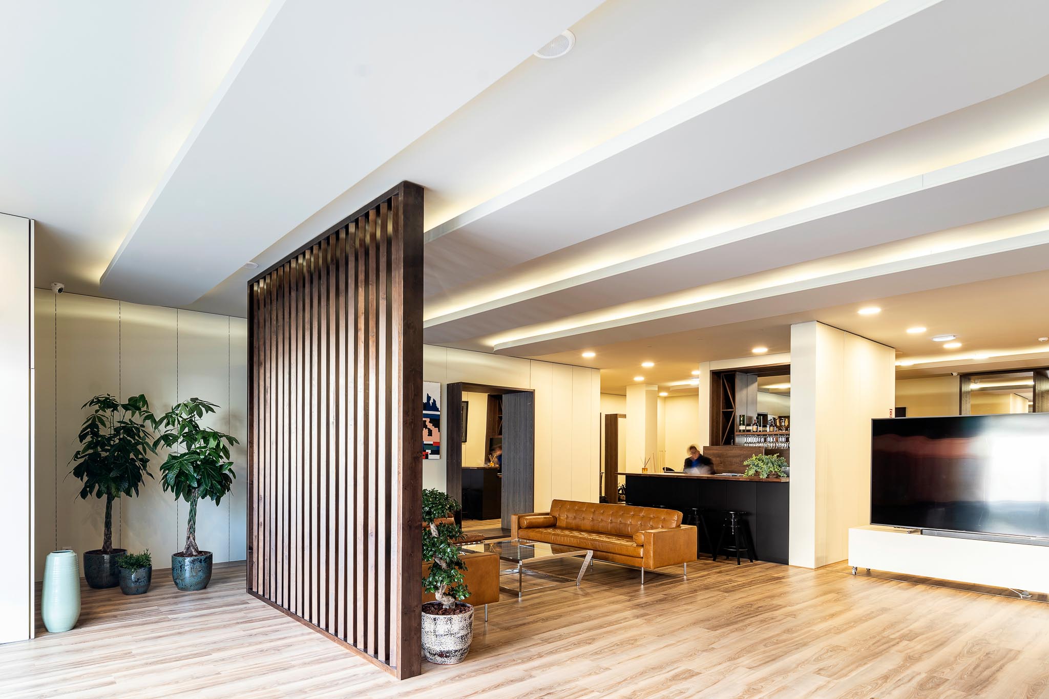 Sala moderna com sofá de couro castanho, mesa de vidro, plantas em vasos e parede divisória de ripas de madeira.