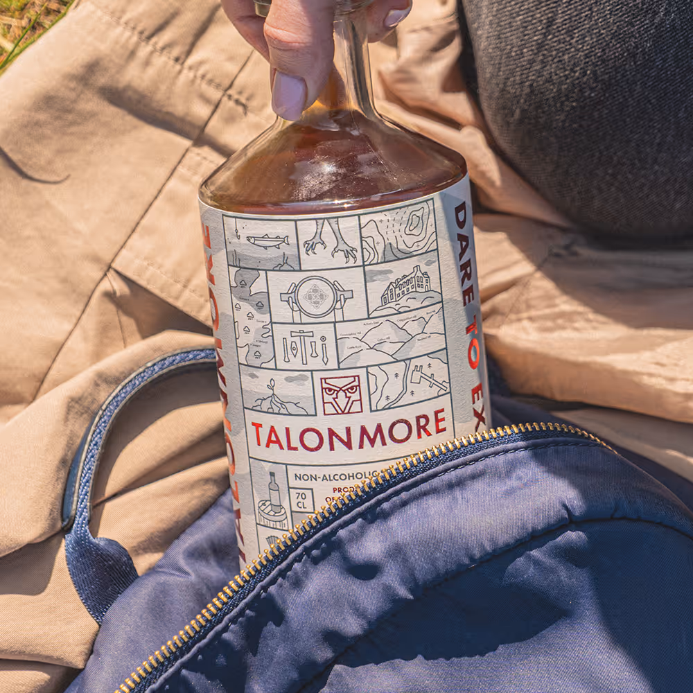 Hand placing a Talonmore non-alcoholic spirit bottle into a partially open blue backpack on a beige fabric surface.