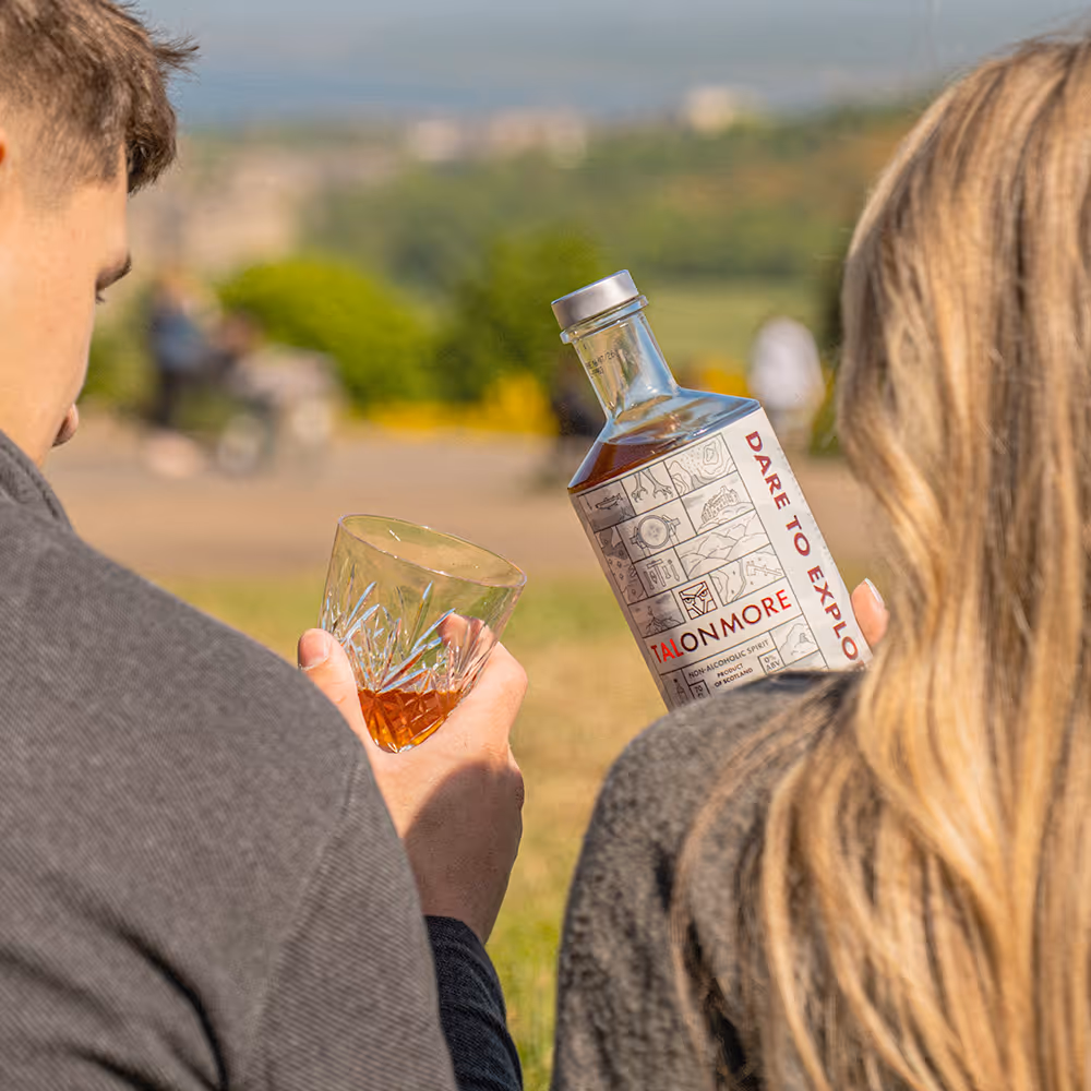 Two people holding a crystal glass with amber liquid and a bottle labeled Talonmore non-alcoholic spirit outdoors.