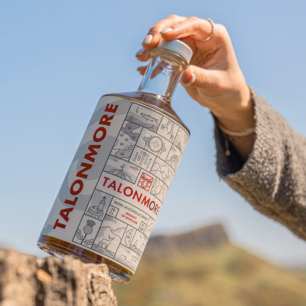 Hand holding a bottle of Talonmore non-alcoholic spirit with a scenic blurred landscape in the background.