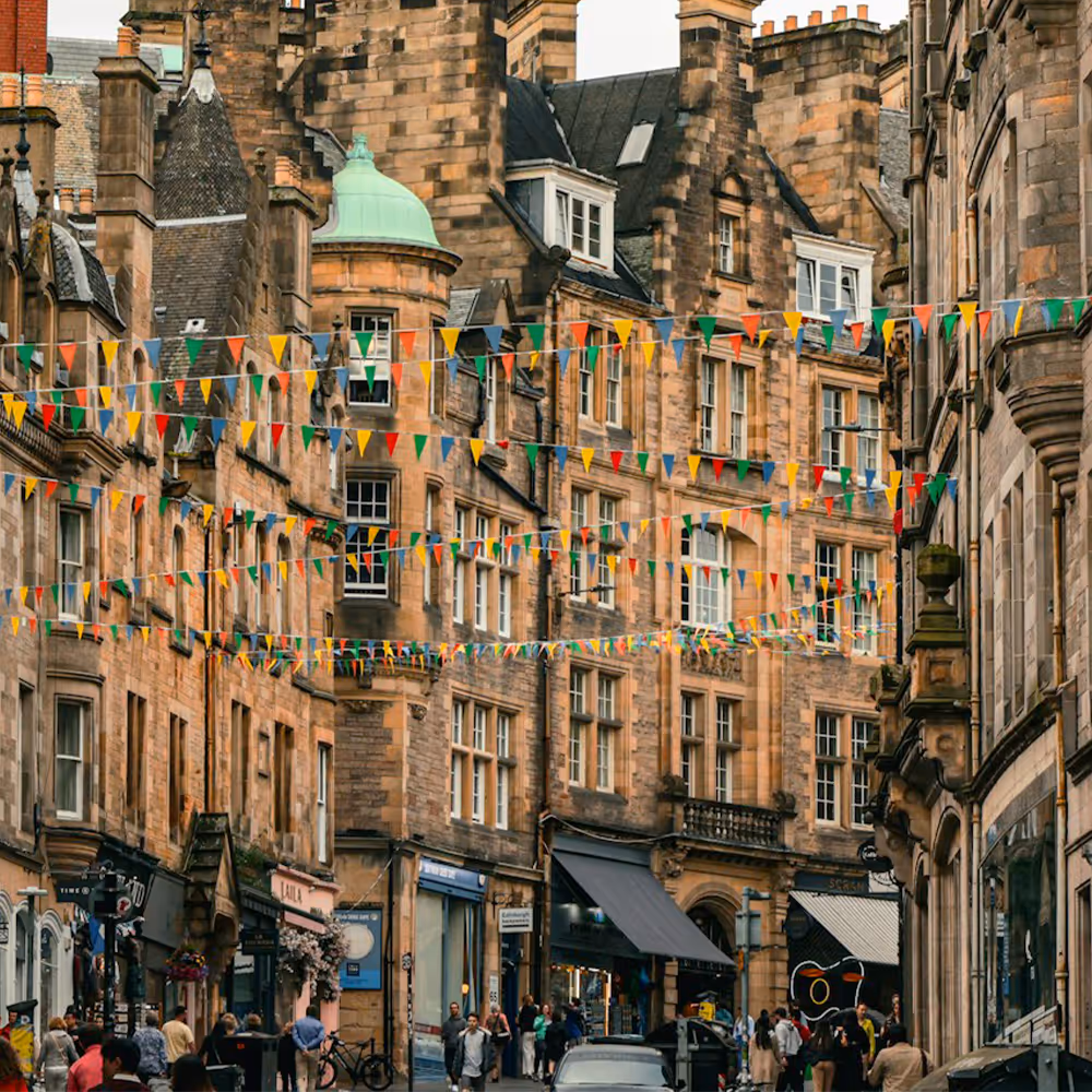 Street scene in a historic city with tall stone buildings adorned with colorful triangular festival bunting and pedestrians walking along the sidewalk.
